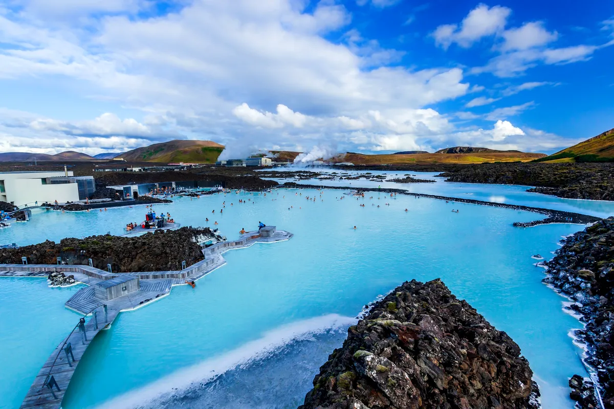 The iconic Blue Lagoon in Iceland, featuring crystal-clear blue waters surrounded by volcanic rocks under a bright, partly cloudy sky.