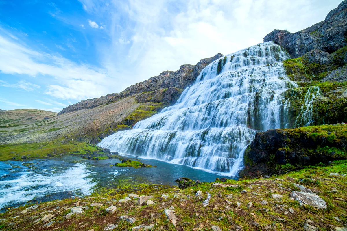 A breathtaking view of Dynjandi Waterfall, cascading down multiple tiers over a rocky slope in the Westfjords of Iceland. The vibrant green landscape adds a striking contrast to the pure white water as it flows into the river below. The blue skies enhance the sense of serenity and natural beauty.