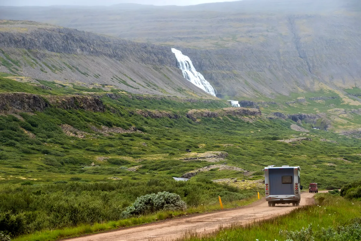 An RV travels along a gravel road towards Dynjandi Waterfall, visible in the distance cascading down the mountainside. The lush green landscape of the Westfjords stretches out on either side, providing a serene and peaceful setting for this journey through Iceland's remote wilderness.