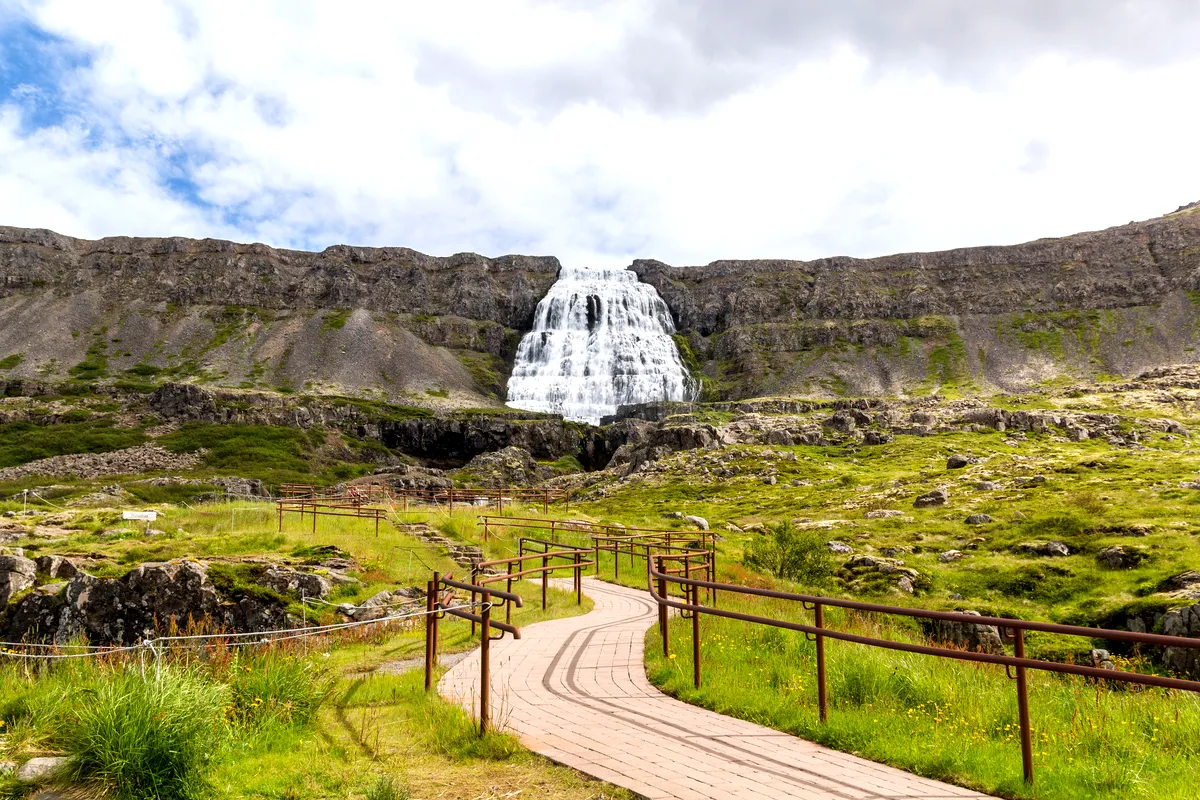 A paved hiking path winds through a green valley leading towards the majestic Dynjandi Waterfall in the Westfjords of Iceland. The towering waterfall is visible in the distance, creating a serene and inviting scene for hikers and nature lovers.