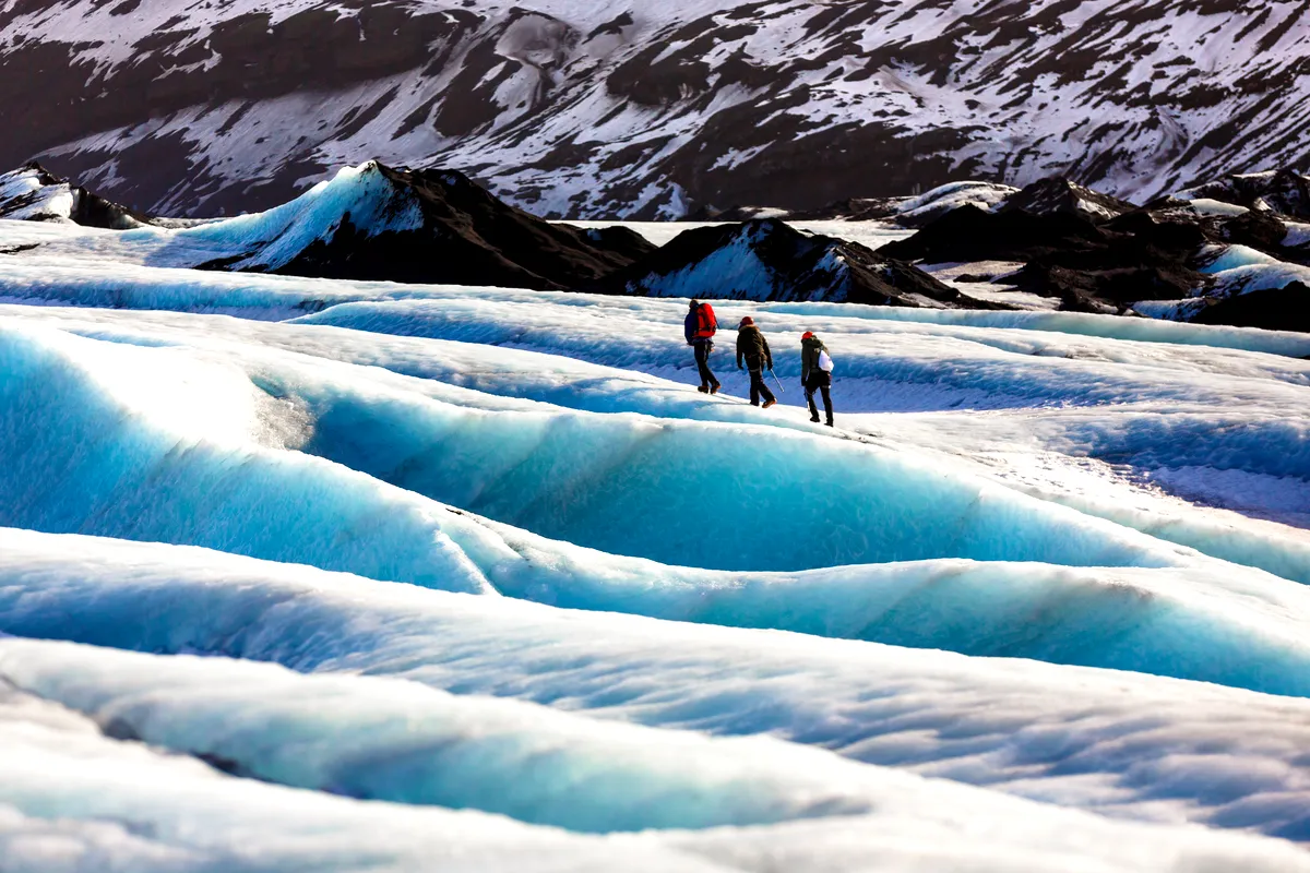 Three hikers traverse the brilliant blue ice of a glacier in Iceland, surrounded by dark volcanic rock and snow-covered mountains.