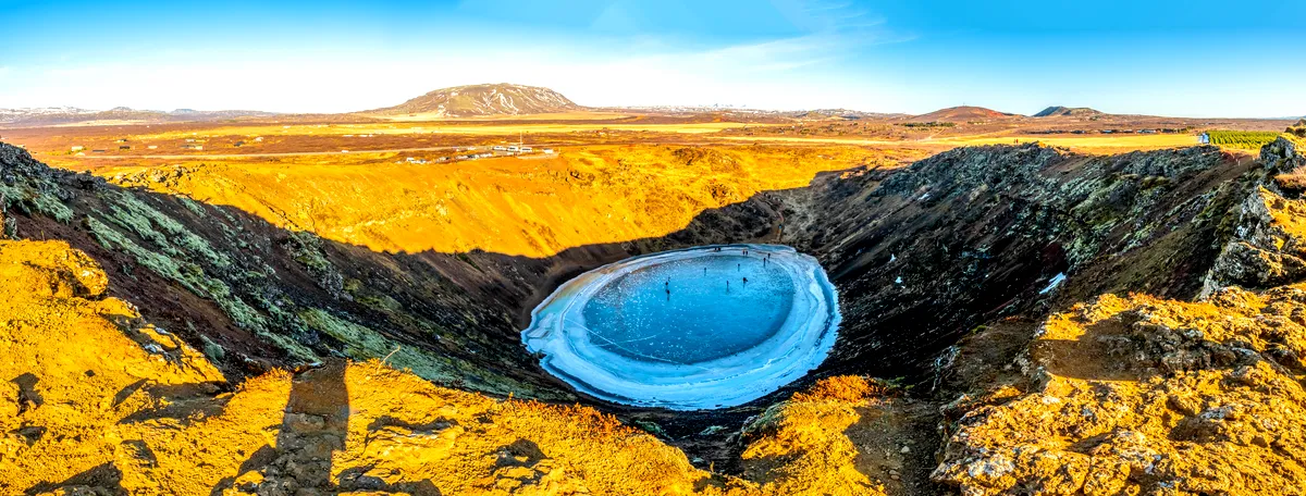 A panoramic view of a volcanic crater in Iceland with a frozen lake at the bottom. The crater's steep walls are covered in rugged rock formations, with the surrounding landscape bathed in warm golden hues under a clear blue sky. The scene highlights the stark and dramatic beauty of Iceland's volcanic terrain during March.