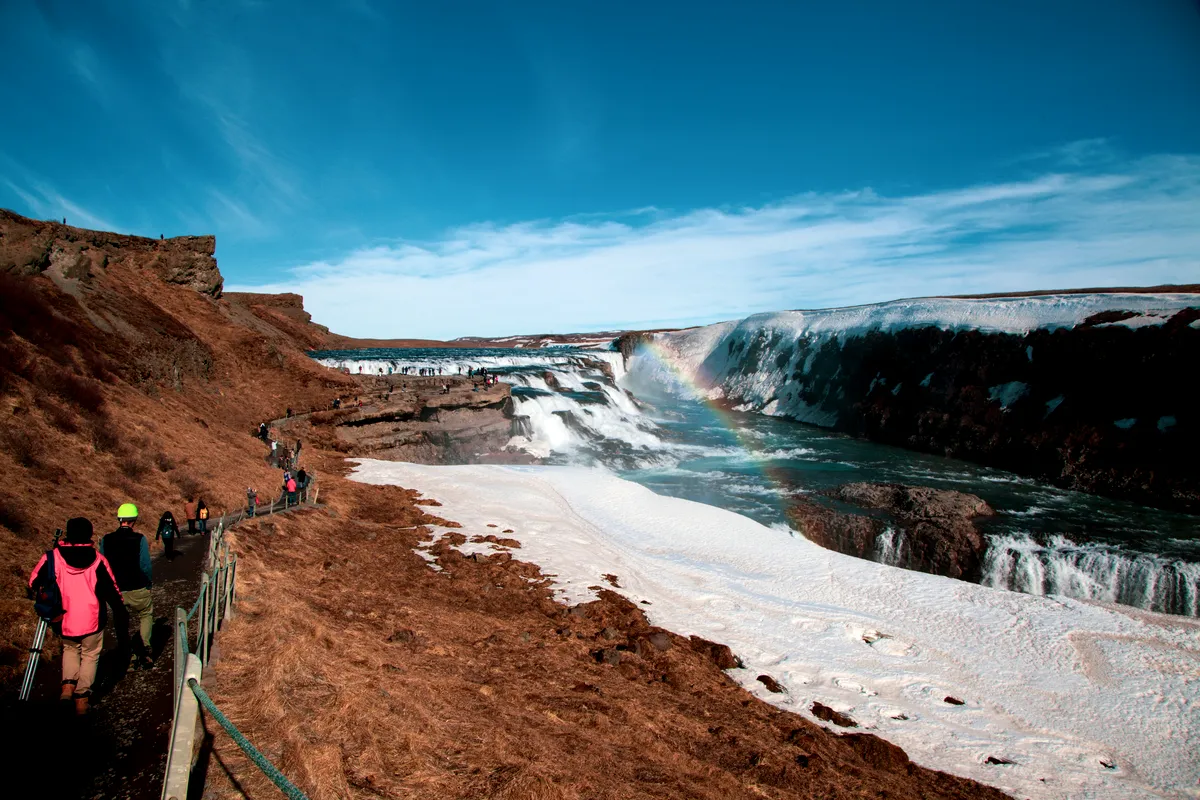The iconic Gullfoss waterfall flows over partially snow-covered terrain. A vibrant rainbow arches across the waterfall as visitors walk along the path, capturing the magical blend of water, ice, and light.