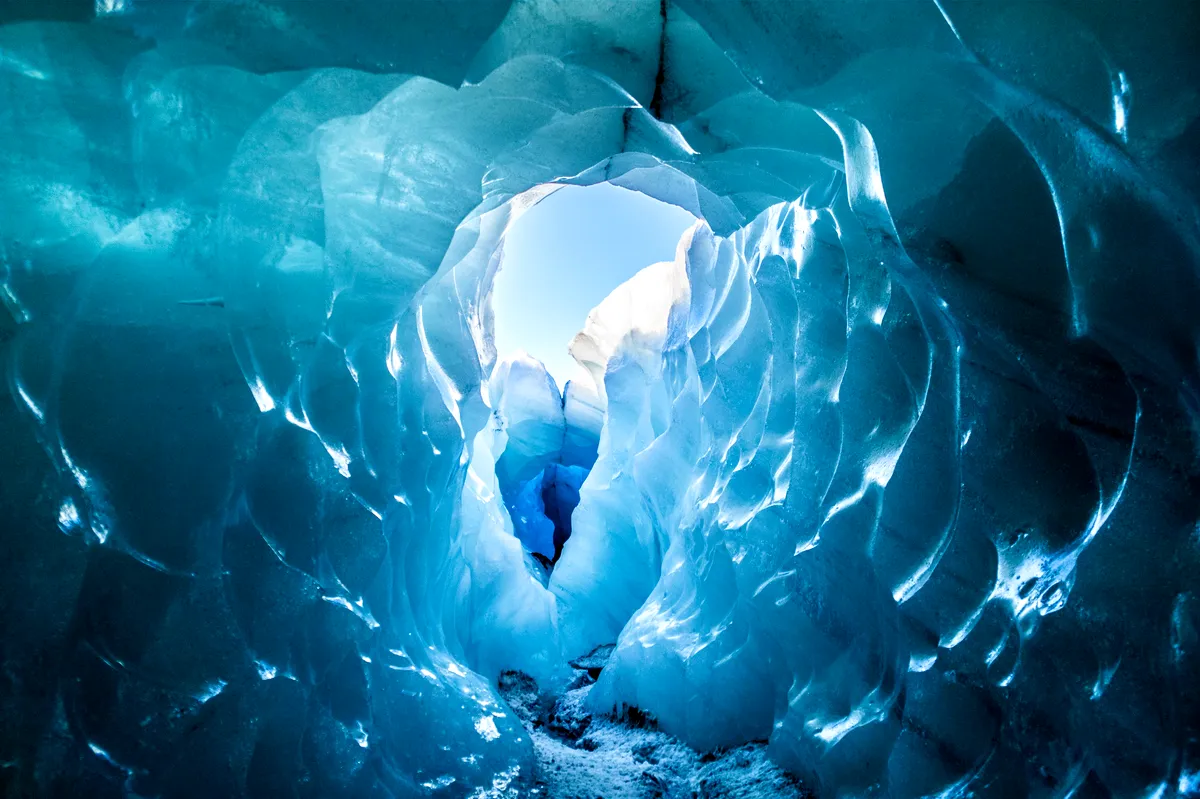 A mesmerizing view from inside an ice cave in Iceland. The icy walls are illuminated in various shades of blue, creating a tunnel-like formation leading to the outside. The ice appears sculpted and layered, with light filtering through, enhancing the vibrant blue hues. The opening at the end of the cave reveals a glimpse of the bright sky and the snowy landscape beyond, adding a contrast between the deep blue interior and the bright, sunlit exterior.