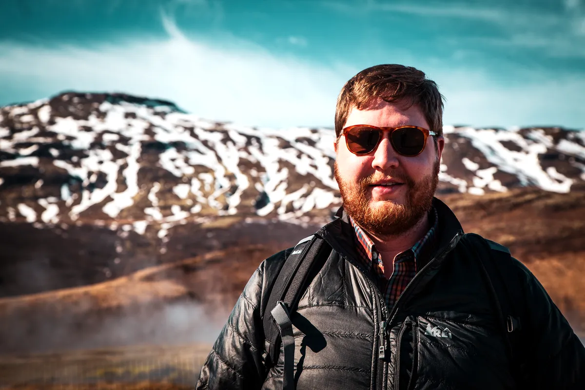 A man in a black jacket and sunglasses stands against a mountainous backdrop with snow patches, exemplifying the cool yet pleasant weather of Iceland in April.