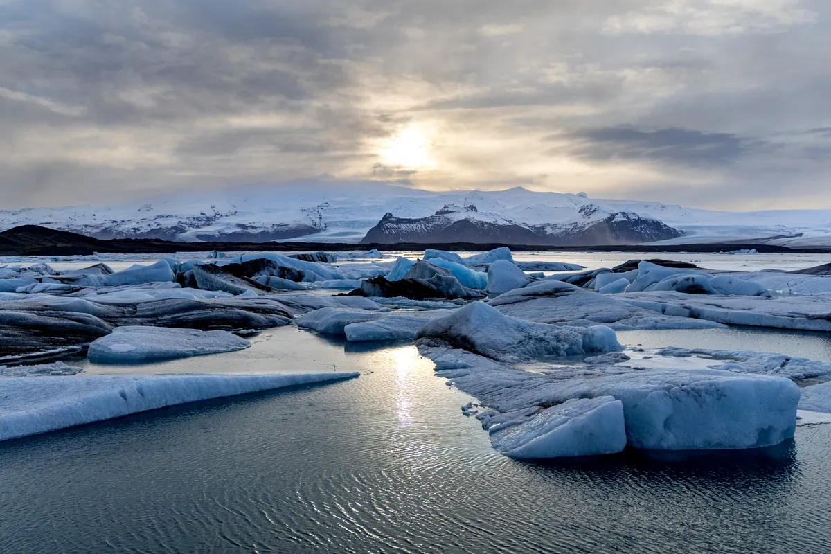 Massive chunks of glacial ice float peacefully in the still waters, reflecting the soft light of the Arctic sun. Snowy peaks loom in the background, adding to the pristine beauty of Iceland’s frozen landscape.