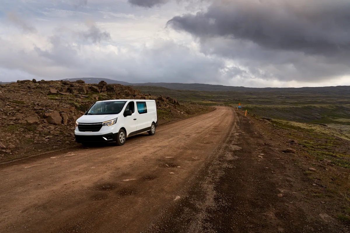 A white campervan is parked on a remote, unpaved road in Iceland. The landscape around is rugged, with rocky terrain and distant hills under a dramatic, cloudy sky. The road stretches ahead into the horizon, suggesting a sense of adventure and exploration in the vast, untouched wilderness.