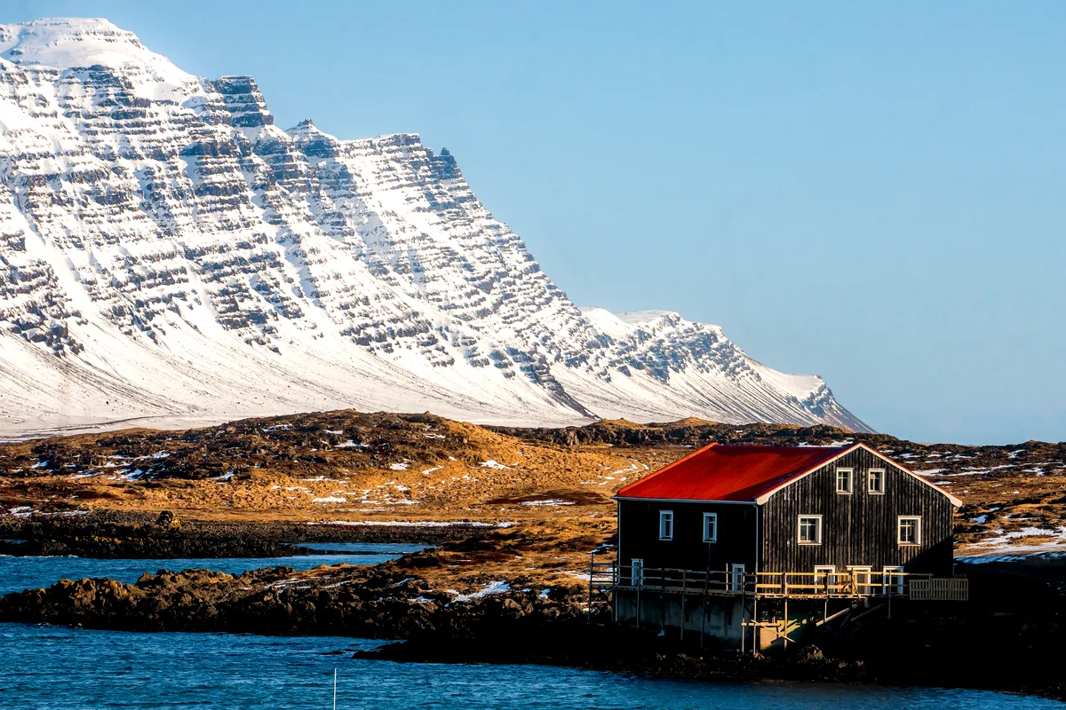 A rustic black wooden cabin with a red roof sits on the edge of a rocky shoreline in Iceland, overlooking the calm blue waters of a fjord. In the background, a massive snow-covered mountain range with intricate layers of rock rises majestically under a clear blue sky, creating a stunning contrast between the rugged terrain and the peaceful setting of the cabin.