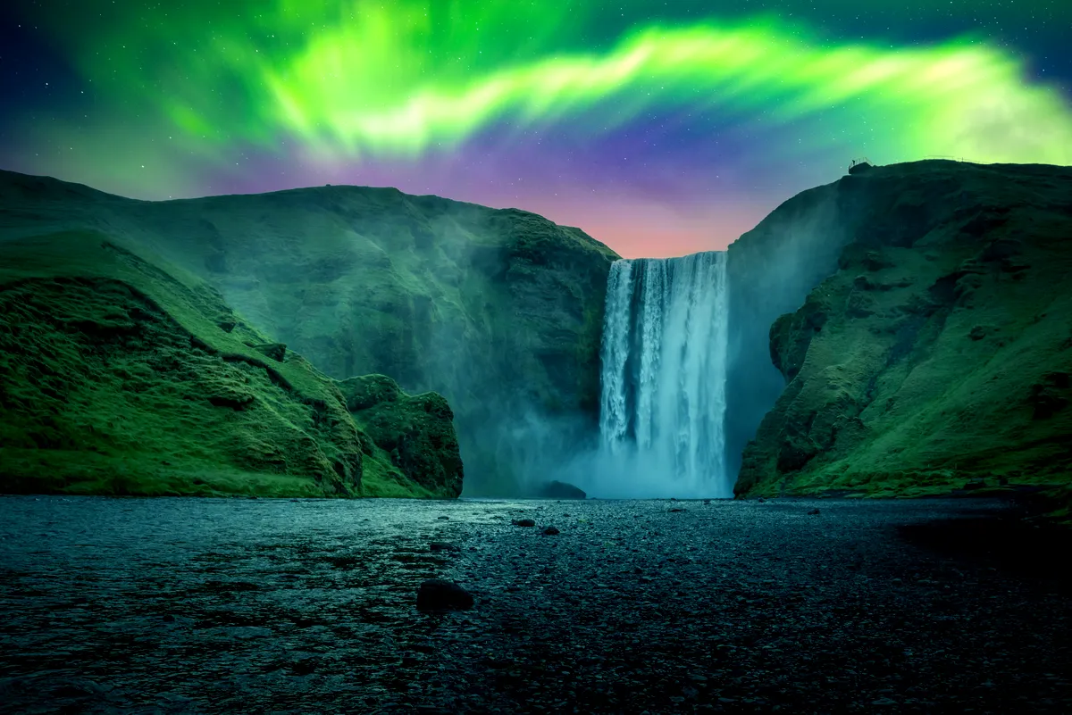 Vibrant Northern Lights over Skógafoss waterfall in Iceland, with the sky glowing in green and purple hues against the dramatic cascade of water.