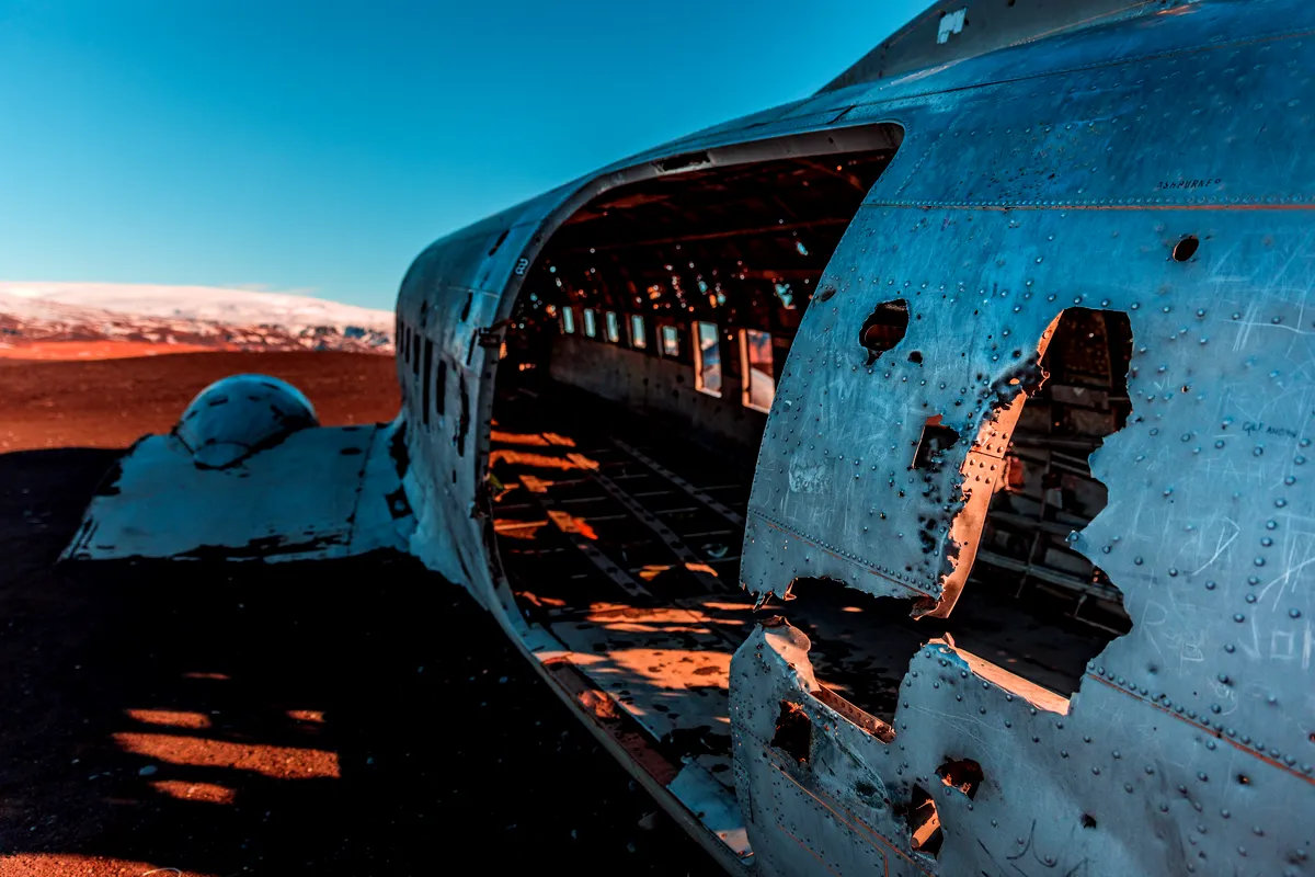 A close-up view of the US Navy DC-3 plane wreck on Sólheimasandur beach, Iceland. The metallic body of the plane is rusted and corroded, with large holes in the fuselage. The interior is exposed, showing the ribbed structure of the cabin. The warm sunlight highlights parts of the plane, contrasting against the cold, barren black sand and distant snow-covered mountains under a clear blue sky.