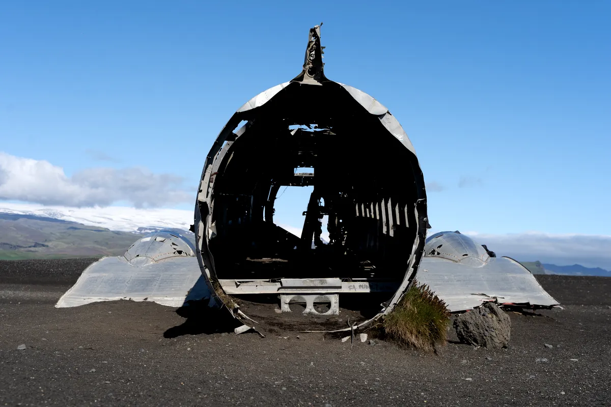 A head-on view of the wreckage of the US Navy DC-3 plane on the black sand beach of Sólheimasandur, Iceland. The image shows the hollow, cylindrical fuselage with no cockpit or rear, creating a tunnel-like appearance. The barren, dark sand surrounds the wreckage, with mountains and a clear blue sky visible in the background. The rusted and worn remains of the plane contrast sharply against the natural, desolate landscape.