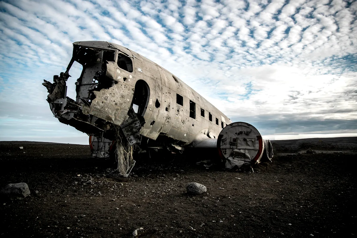 The wreckage of a US Navy DC-3 airplane at Sólheimasandur beach in Iceland, under a sky filled with rippling clouds. The plane, weathered and corroded, lies in stark contrast to the desolate, black sand landscape. The cockpit is heavily damaged, with parts of the fuselage missing. The surrounding area is barren, with only rocks and dark soil visible, adding to the eerie and isolated atmosphere of the scene.