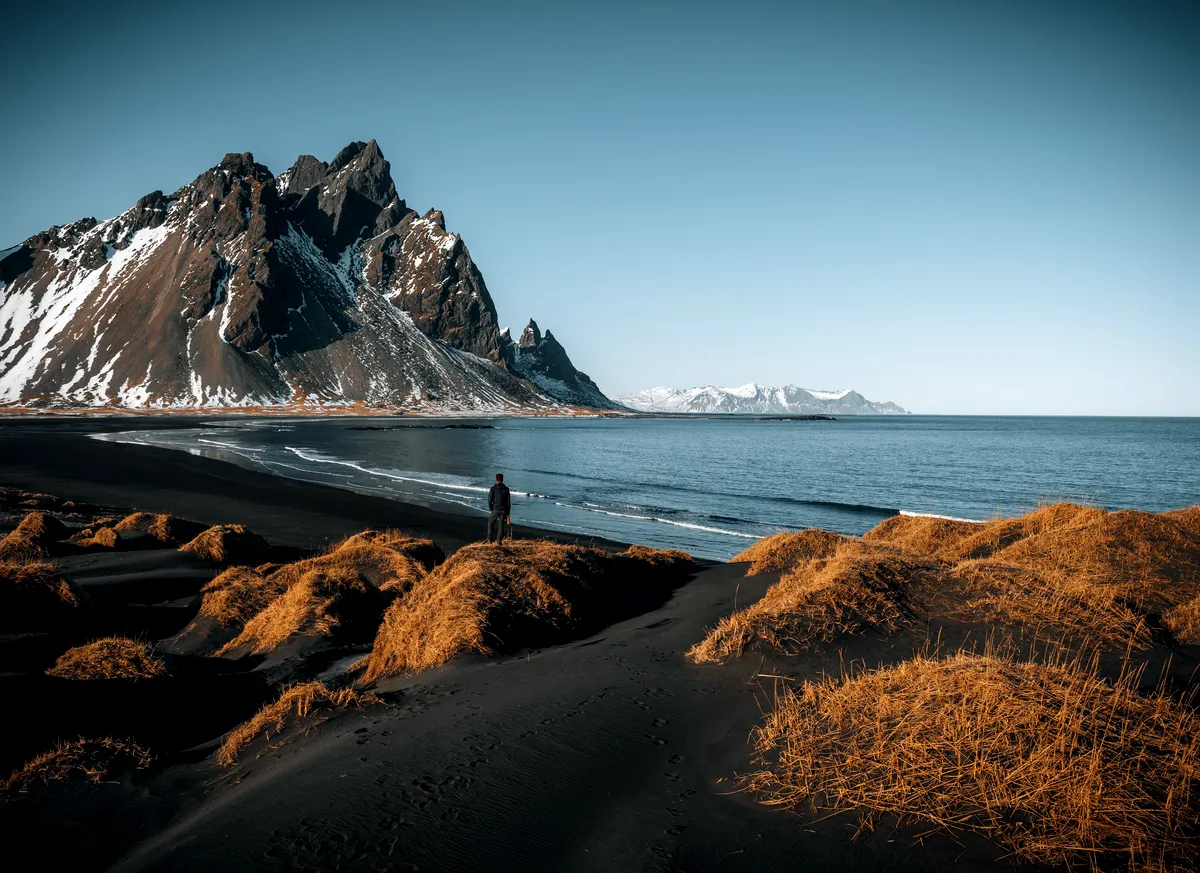 A scenic view of a person standing on a black sand dune, gazing at the towering snow-capped peaks of the Stokksnes mountain range in Iceland. The serene blue ocean stretches out towards the horizon, and the beach is dotted with patches of golden-brown grass. The sky is clear, creating a peaceful and awe-inspiring atmosphere.