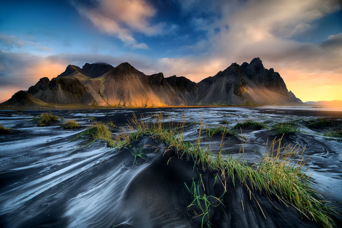A stunning view of the Stokksnes Peninsula in Iceland, featuring dramatic mountains in the background with soft, golden sunlight illuminating their rugged peaks. In the foreground, black sand dunes adorned with patches of green grass create a surreal contrast against the backdrop of the setting sun. The sky is painted in a gradient of blue and orange hues as clouds drift lazily overhead.