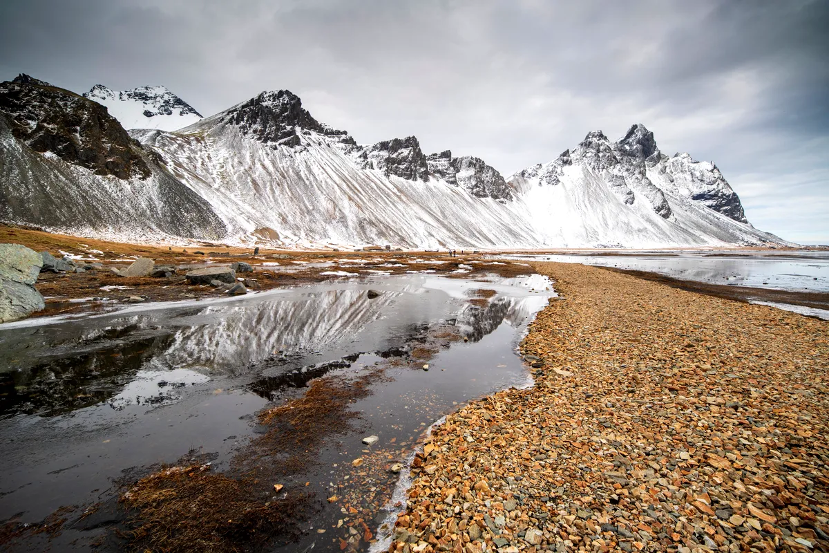 A breathtaking landscape of Vestrahorn Mountain in Iceland, featuring a snow-covered mountain range with sharp peaks reflected in the still water of a puddle on the rocky shoreline. The dark clouds above create a moody atmosphere, enhancing the contrast between the icy mountains and the barren earth below.